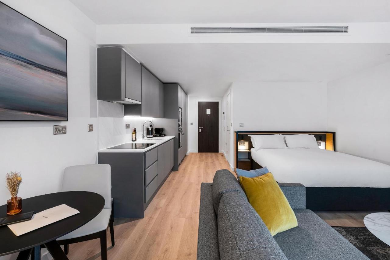 Citysuites 2 Aparthotel Manchester Bagian luar foto The photo shows a modern and stylish studio apartment. It features a kitchenette with dark cabinetry and a small dining table with two chairs. There's a cozy sofa with colorful cushions, positioned next to the table. In the background, a large bed wi