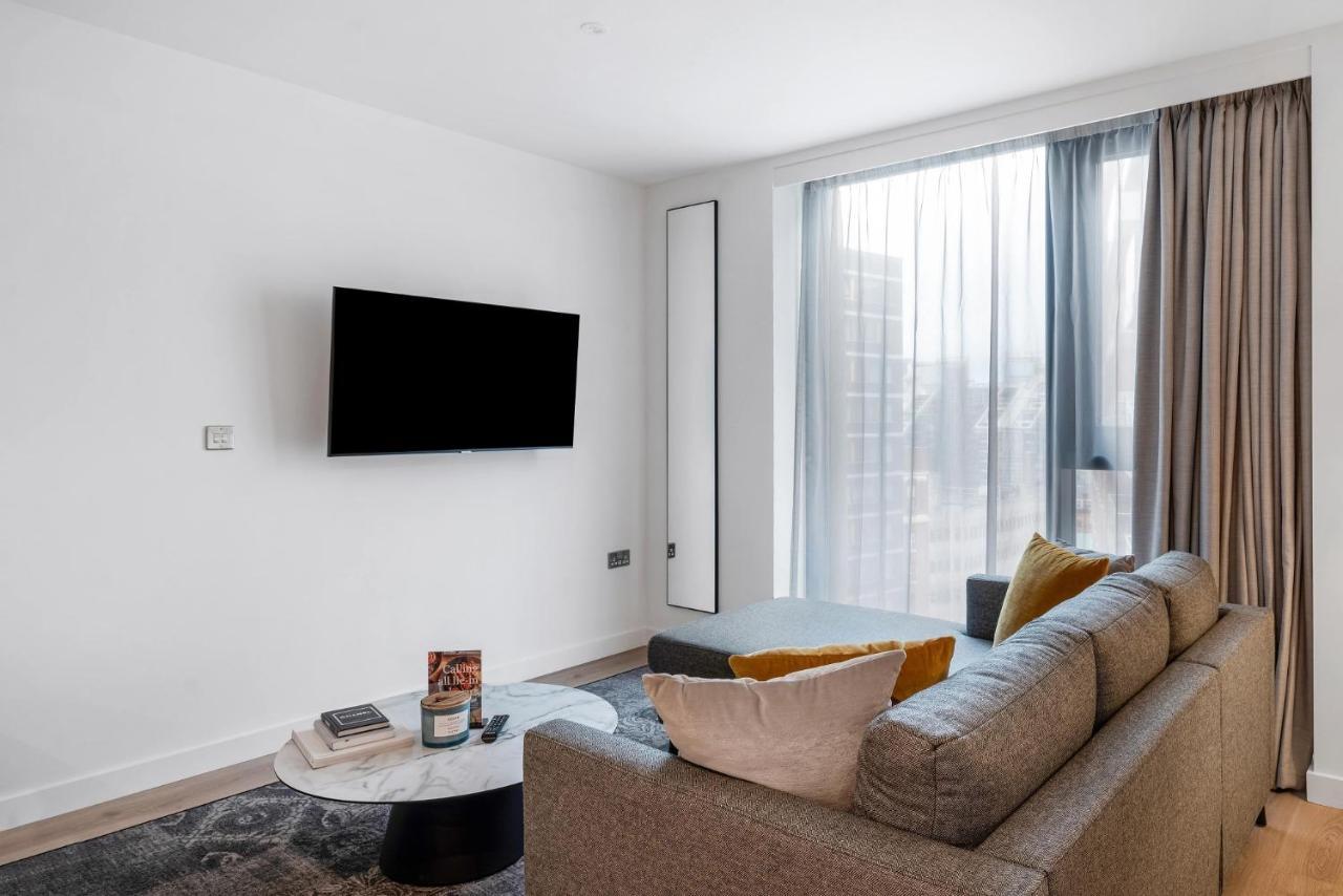 Citysuites 2 Aparthotel Manchester Bagian luar foto The image shows a modern living room with a minimalist design. There is a large flat-screen TV mounted on the white wall. A comfortable gray sofa with textured cushions occupies the center of the room, complemented by a round coffee table in front of