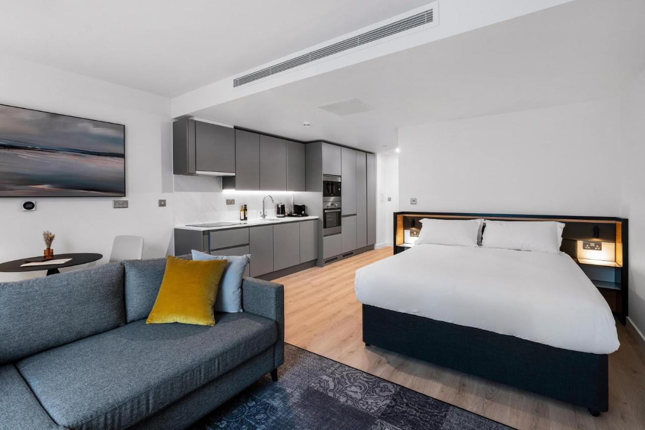 Citysuites 2 Aparthotel Manchester Bagian luar foto The photo shows a modern studio apartment. On one side, there is a comfortable gray sofa adorned with a yellow cushion. Nearby, a round table is placed, likely intended for dining or casual sitting. 

The kitchen area features sleek gray cabinetry wi