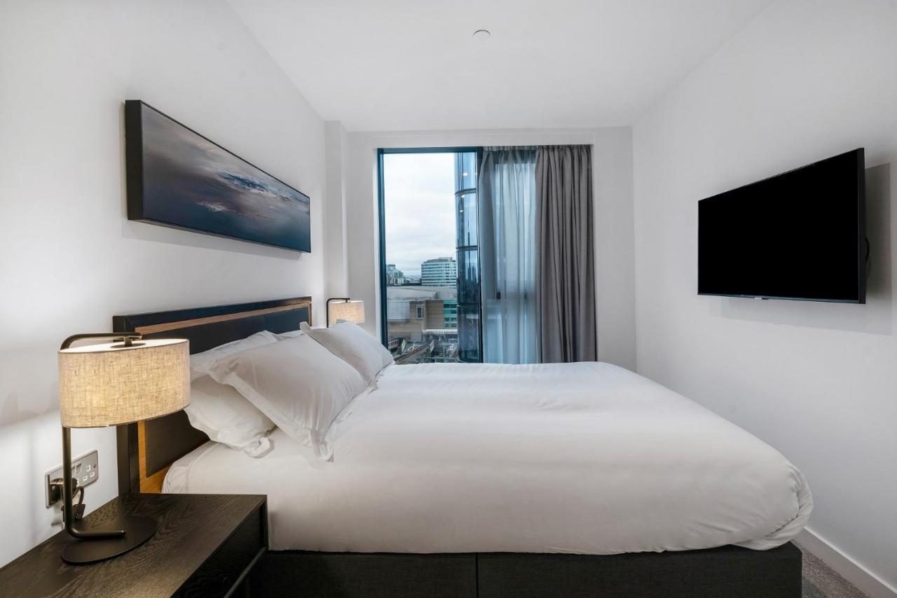 Citysuites 2 Aparthotel Manchester Bagian luar foto The photo shows a modern bedroom with a minimalist aesthetic. It features a large bed with white bedding and two pillows. On either side of the bed, there are bedside tables with lamps. A large window lets in natural light, covered by sheer curtains.