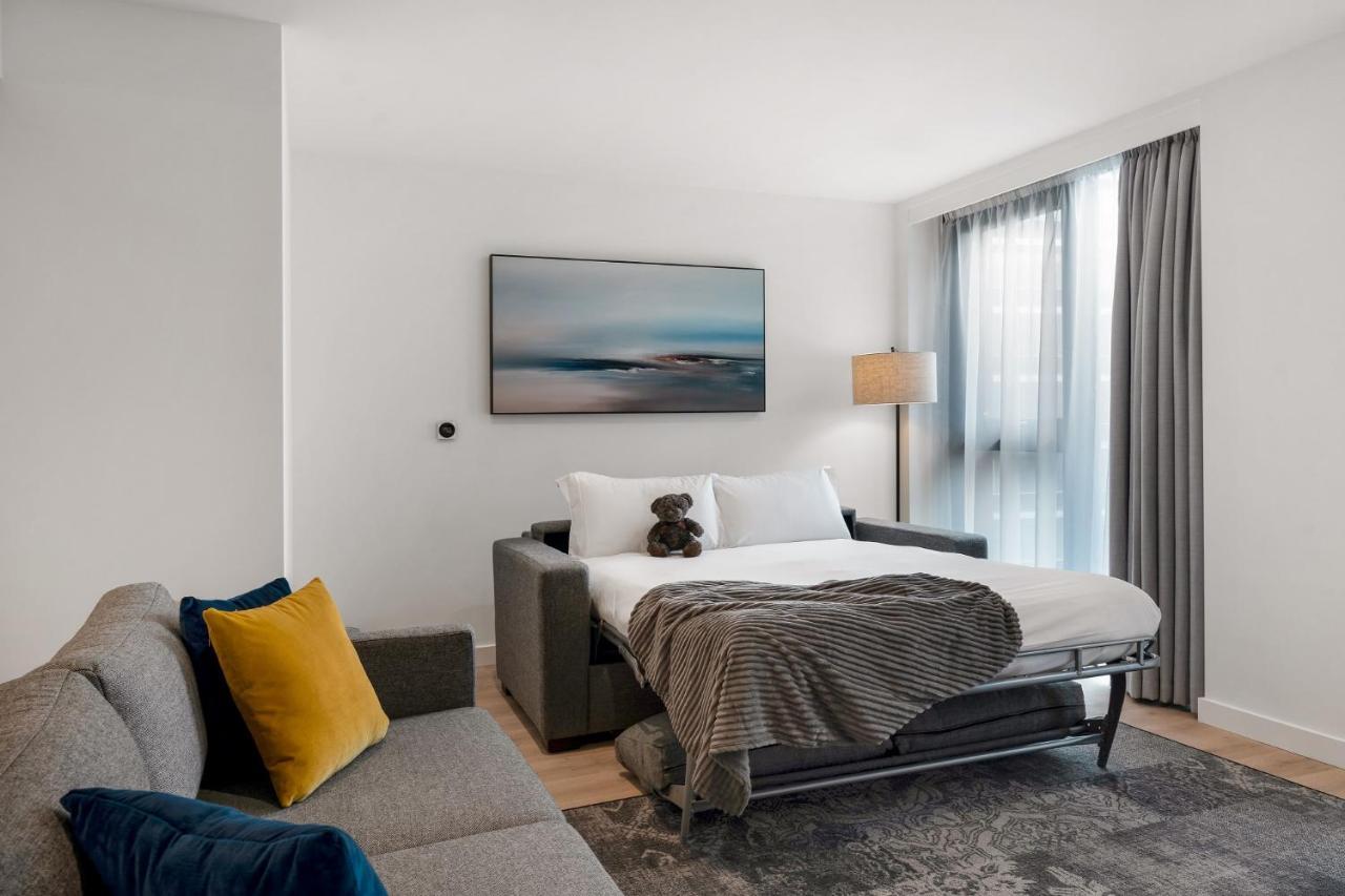 Citysuites 2 Aparthotel Manchester Bagian luar foto The photo shows a modern and minimalistic guest room. There is a sofa on the left side, adorned with a colorful cushion. In the middle is a bed that appears to be a pull-out style, featuring white bedding and a gray throw blanket. A stuffed toy is pl