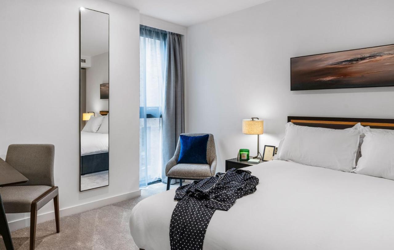 Citysuites 2 Aparthotel Manchester Bagian luar foto The photo shows a modern hotel room. It features a neatly made bed with white linens and a dark-colored throw draped over it. There is a small bedside table with a lamp next to the bed. A comfortable chair in a light color is positioned near the wind