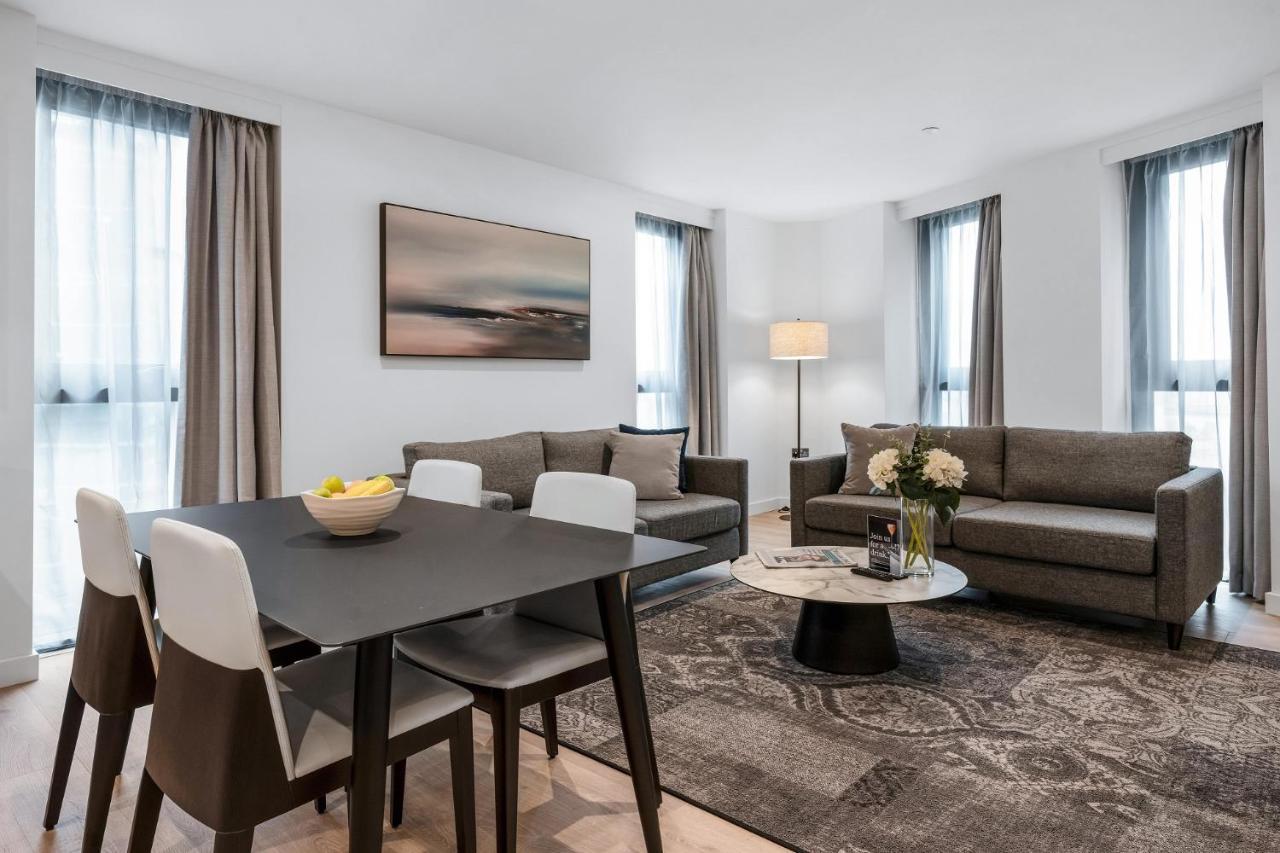 Citysuites 2 Aparthotel Manchester Bagian luar foto The photo depicts a modern and stylish interior of a living space. On the left, there is a dining table with four chairs, featuring a bowl of fruits on the table. In the background, there's a cozy seating area with a gray sofa and a smaller round cof