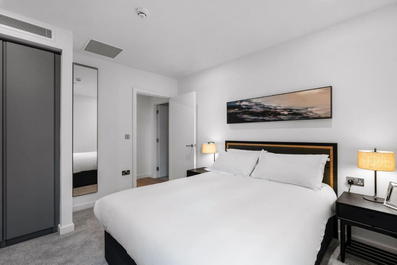 Citysuites 2 Aparthotel Manchester Bagian luar foto The photo shows a modern bedroom featuring a large double bed with white bedding. There are two bedside tables with lamps on either side of the bed. The walls are painted white, and there's a piece of artwork above the bed that depicts a landscape. A