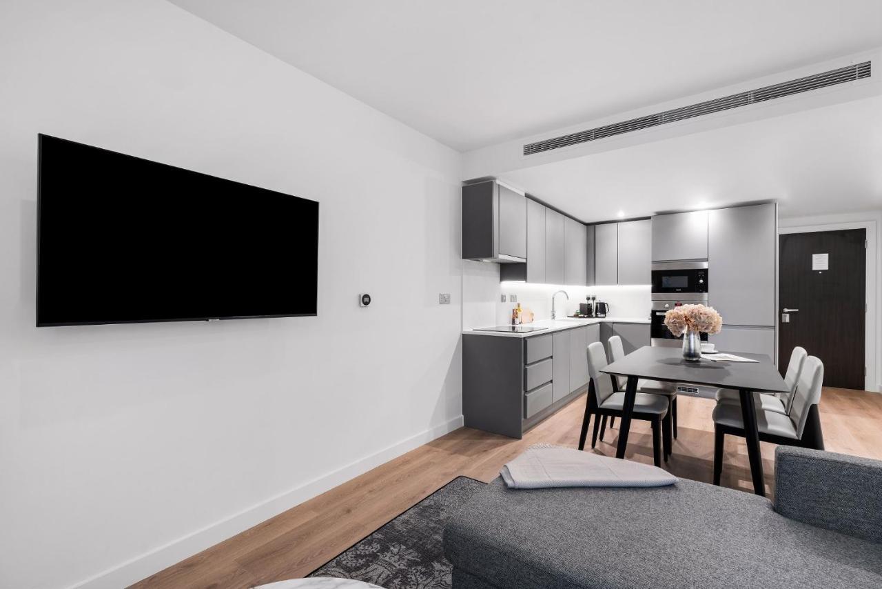 Citysuites 2 Aparthotel Manchester Bagian luar foto The photo shows a modern, minimalist living space. On the left, there is a large flat-screen TV mounted on the wall. To the right, a kitchen area features sleek gray cabinets and appliances, including a built-in microwave. In front of the kitchen, th