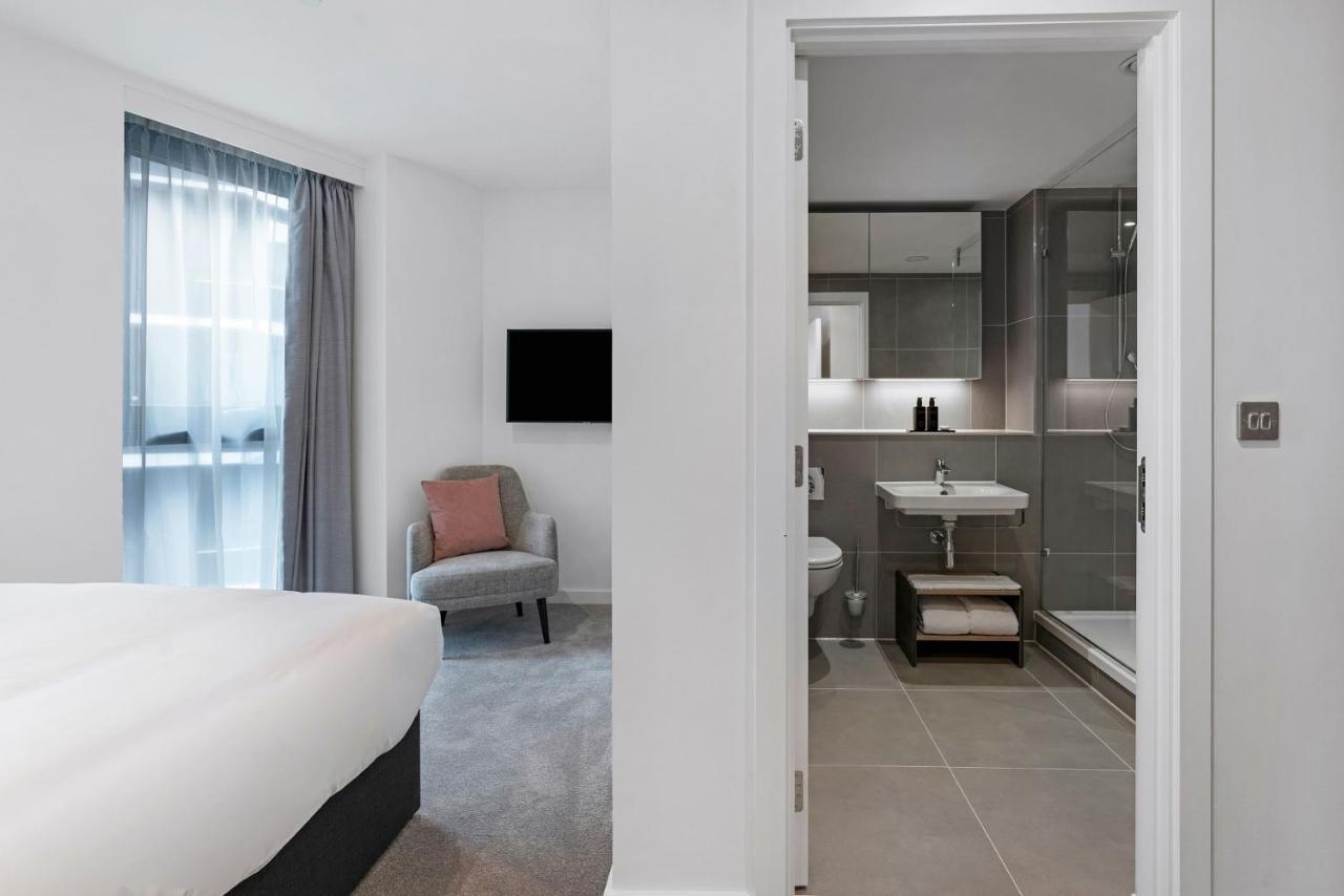 Citysuites 2 Aparthotel Manchester Bagian luar foto The photo shows a modern hotel room and bathroom setup. On the left, there is a bed with white bedding and a small sitting area that includes a gray chair and a pillow in a subtle pink shade. A flat-screen TV is mounted on the wall, providing enterta