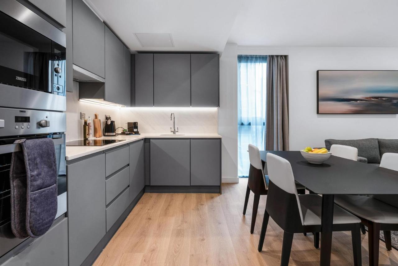 Citysuites 2 Aparthotel Manchester Bagian luar foto The photo shows a modern kitchen and dining area. The kitchen features sleek gray cabinetry, a white countertop, and built-in appliances such as an oven. There is a sink and some kitchen utensils displayed. The adjacent dining area contains a black t
