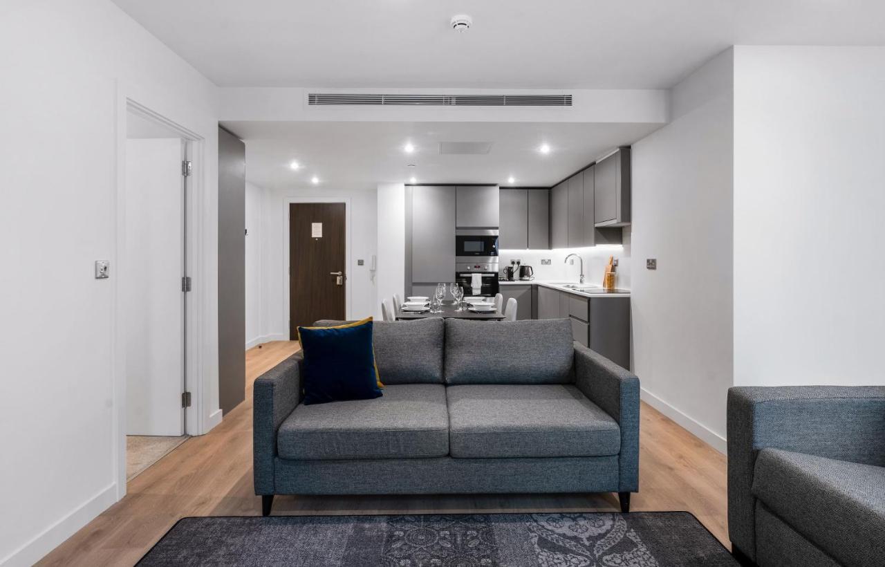 Citysuites 2 Aparthotel Manchester Bagian luar foto The photo shows a modern interior space, likely a living area or apartment. In the foreground, there is a gray sofa with a blue cushion positioned on a textured rug. To the back, there is an open kitchen with sleek cabinetry and appliances, visible a