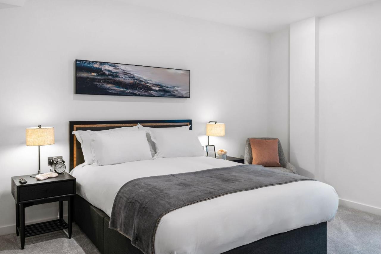 Citysuites 2 Aparthotel Manchester Bagian luar foto The photo shows a modern bedroom featuring a large bed with crisp white linens and a grey throw blanket. On either side of the bed, there are bedside tables with lamps. A piece of abstract art hangs above the headboard, and there is a chair in a corn