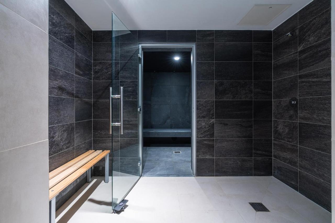 Citysuites 2 Aparthotel Manchester Bagian luar foto The photo shows a modern, minimalist shower area. The space is predominantly tiled with dark gray tiles on the walls, giving it a sleek and sophisticated look. There is a glass door that leads to another room, and a wooden bench is positioned against