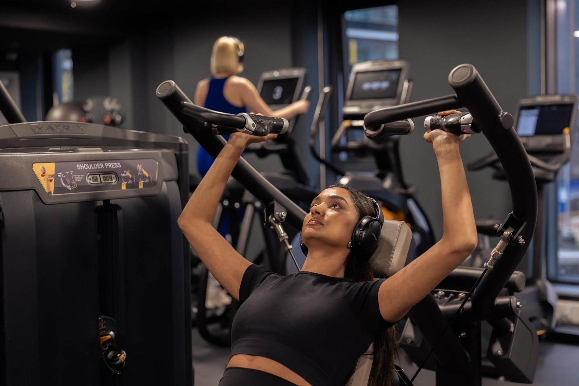 Citysuites 2 Aparthotel Manchester Bagian luar foto The photo shows a gym setting with two individuals engaged in physical exercise. In the foreground, a woman is using a chest press machine while wearing headphones, focusing on her workout. She appears to be in a seated position, pushing the handles 