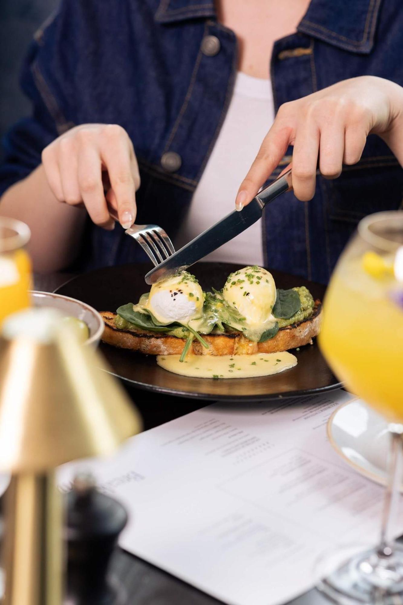 Citysuites 2 Aparthotel Manchester Bagian luar foto The photo shows a person preparing to eat a dish of eggs Benedict. They are holding a knife and fork, ready to cut into the meal, which features poached eggs on top of a slice of toast, accompanied by spinach and a creamy hollandaise sauce drizzled o