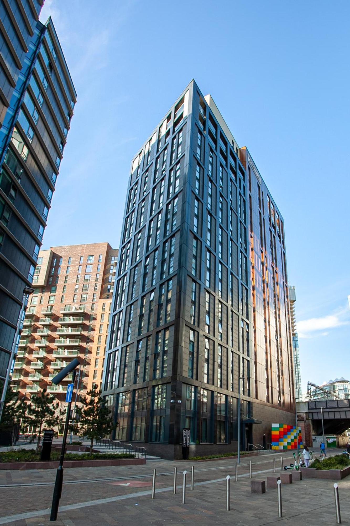 Citysuites 2 Aparthotel Manchester Bagian luar foto The photo shows a modern high-rise building with a sleek, glass façade. It is set against a blue sky, indicating a clear day. The building appears to have multiple floors with large windows that reflect light. Nearby, there are additional buildings, 